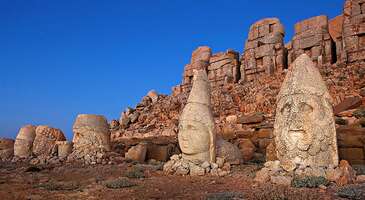 Adıyaman&rsquo;da konumlanan efsane dağ, resmiyette Nemrut Dağı Milli Parkı olarak anılıyor. Dolayısıyla Bitlis-Tatvan&rsquo;da bulunan Nemrut Dağı ile karıştırılmaması gerekiyor. UNESCO D&uuml;nya Mirası Listesi&rsquo;nde de yer alan Nemrut Dağı Milli Parkı, Anadolu&rsquo;da g&ouml;r&uuml;lmesi gereken yerlerin başında geliyor. Toros Dağları'nın doğusundaki en y&uuml;ksek noktalardan biri olan ve 2.150 metrede konumlanan Nemrut Dağı&rsquo;nı keşfetmeye hazırsan arkeolojik yapıları, efsanevi heykelleri ve daha niceleriyle dolu bir yolculuğa &ccedil;ıkıyoruz! Nemrut Dağı&rsquo;nın hikayesi Nemrut Dağı&rsquo;nın ve i&ccedil;erisinde bulunan &ouml;ren yerinin ilk keşfedilişi 1881 yılında ger&ccedil;ekleşiyor. Bu tarihi hazineyi keşfeden kişi, o sıra y&ouml;rede g&ouml;revli olan Alman m&uuml;hendis Karl Sester&rsquo;dir. Bir gezinti esnasında tesad&uuml;fen Nemrut Dağı&rsquo;nın meşhur heykelleriyle karşılaşıyor. Harabelerde, tanrı heykellerinin oturtulduğu kaidelerin &uuml;zerinde Grek&ccedil;e yazıtlar olmadığı i&ccedil;in Asur d&ouml;nemine ait olduklarını d&uuml;ş&uuml;nerek İzmir&rsquo;deki Alman konsolosluğuna haber verir. 1882 yılına gelindiğinde Karl Sester ve Otto Puchstein b&ouml;lgede incelemeler yapmaya başlar. Ardından 1883 yılında M&uuml;ze-i H&uuml;mayun&rsquo;un m&uuml;d&uuml;r&uuml; Osman Hamdi Bey de ekibiyle birlikte bu &ccedil;alışmaya katılır. Ayrıca II. D&uuml;nya Savaşı sonrası &uuml;nl&uuml; arkeologlardan Theresa Goell ve Alman Karl Doerner de Nemrut&rsquo;ta birtakım araştırma ve inceleme faaliyetleri y&uuml;r&uuml;t&uuml;r. T&uuml;m bu &ccedil;alışmalar neticesinde b&ouml;lgedeki harabe ve devasa tanrı heykellerinin Asur Medeniyeti&rsquo;ne değil Kommagene Krallığı&rsquo;na ait olduğu ve M.&Ouml; 163 &ndash; M.S 72 tarihleri arasında yaptırıldıkları tespit edilir. Bu değerli yapıların iki bin yıl gibi uzun bir s&uuml;re kaderine terk edildiği anlaşılır. Helenistik d&ouml;nemin en g&uuml;zel &ouml;rneklerinden olan anıtsal heykeller ve mezarlar Kommagene Kralı I. Antiochos tarafından yaptırılır. 2150 m y&uuml;ksekliğindeki Nemrut Dağı&rsquo;nın yama&ccedil;larında yer alan bu heykel ve mezarlar, atalara ve tanrılara olan minnettarlığın bir nişanesi olarak yerini alır. Değerli bir &ouml;ren yeri olduğu anlaşılan Nemrut Dağı, 11 Aralık 1987&rsquo;de UNESCO D&uuml;nya Mirasları Listesi&rsquo;ne dahil edilir. UNESCO D&uuml;nya Mirası Listesi'ne giren diğer tarihi ve doğal g&uuml;zelliklerimizi keşfetmek i&ccedil;in linke tıklayabilirsin. Nemrut Dağı'nda gezilecek ve g&ouml;r&uuml;lecek yerler Nemrut Dağı Milli Parkı i&ccedil;erisinde yer alan anıtsal heykel ve yapılar, doğu, batı ve kuzey teraslarına yayılıyor. &Ouml;zellikle doğu terası, daha merkezi ve kutsal kabul ediliyor. &Ccedil;&uuml;nk&uuml; &ouml;nemli kabul edilen mimari yapı ve heykel kalıntıları burada yer alıyor. Kire&ccedil;taşı bloklarından yapılan ve g&uuml;n&uuml;m&uuml;zde iyi durumda olan devasa heykellerin boyları 8 &ndash; 10 metre arasında değişiyor. Ayrıca alanda kral mezarının da olduğu d&uuml;ş&uuml;n&uuml;l&uuml;yor. Fakat hen&uuml;z nerede olduğu bilinmiyor. Bunlara ek olarak Nemrut&rsquo;un kitabesinde yazanlar da insanların olduk&ccedil;a ilgisini &ccedil;ekiyor: &lsquo;&lsquo;Ata h&uuml;k&uuml;mdarlığını devraldığım zaman, dindarlığımın bir sonucu olarak, tahtıma bağlı krallığı t&uuml;m tanrıların ortak yurdu yaptım. Zamanın akışı i&ccedil;inde her kim, bu kanunu ve bize ibadeti korur ve s&uuml;rd&uuml;r&uuml;rse, benim hayır dualarımla anılacaktır. T&uuml;m rahmetli atalar ve tanrılar ondan razı olsun. Her kim ki, bu d&uuml;zenin kutsal ge&ccedil;erliliğini bozar ya da zarar verir ya da ger&ccedil;ek anlamını değiştirmeye yeltenirse, yalnız kendisi değil, aynı zamanda t&uuml;m soyu sopu rahmetli atalarımın ve t&uuml;m tanrıların hışmına uğrasın.&rsquo;&rsquo; {insightBoxStart}Yazar Notu:&nbsp;6 Şubat 2023 tarihinde ger&ccedil;ekleşen depremlerde Nemrut Dağı'nın bazı yerlerinde hasar oluştu. Kral heykelleri ise depremde herhangi bir hasar almadı.{insightBoxEnd} Nemrut Dağı heykelleri kimleri simgeliyor? Devasa taş bloklar halinde yan yana sergilenen heykellerin kimleri simgelediği elbette en &ccedil;ok merak edilenlerden biri. Heykeller sırasıyla (sağdan sola doğru) Aslan, Kartal, Antiochus, Kommagene, Zeus, Apollon ve Herakles&rsquo;i simgeliyor. Heykellerin arasında bulunan aslan ve kartal heykelleri ise &ccedil;eşitli kaynaklarda koruyucu hayvan heykelleri olarak ge&ccedil;iyor. Aslan Kommagene Krallığı&rsquo;nın yery&uuml;z&uuml;ndeki hakimiyetini, kartal ise g&ouml;ky&uuml;z&uuml; hakimiyetini simgeliyor. Ek olarak &ldquo;kartal&rdquo; Zeus&rsquo;un buyruklarını insanlara ulaştırmak i&ccedil;in kullandığı bir aracı olarak da simgesel anlam taşıyor. Toplamda 4 adet heykel olması gerekirken g&uuml;n&uuml;m&uuml;zde yalnızca ikisi sergilenebiliyor. {calismasaatleri} Nemrut Dağı giriş &uuml;creti M&uuml;zeKart ile &uuml;cretsiz M&uuml;zeKart'sız girişler 10 euro 18 yaş altı ve 65 yaş &uuml;st&uuml; &uuml;cretsiz. Nemrut Dağı ziyaret saatleri Nemrut Dağı&nbsp;nisan ve kasım ayları arasında ziyaret edilebilir. Haftanın yedi g&uuml;n&uuml; 04.00 ile&nbsp;18.30 saatleri arasında misafirlerini ağırlayan milli park kış sezonu boyunca kapalı. {/calismasaatleri} {insightBoxStart}Yazar Notu:&nbsp;Nemrut Dağı aralık, ocak, şubat ve mart aylarında kapalı oluyor. Bu aylarda ziyaret i&ccedil;in m&uuml;ze m&uuml;d&uuml;rl&uuml;ğ&uuml;nden izin alınması gerektiğini belirtelim. Ayrıca Nemrut Dağı ziyareti i&ccedil;in en m&uuml;kemmel zaman nedir diye sorarsan hi&ccedil; ş&uuml;phesiz g&uuml;ndoğumu derim!{insightBoxEnd} Nemrut Dağı nerede? Nemrut Dağı&rsquo;nın konumu olduk&ccedil;a &ouml;nemli bir konu, &ccedil;&uuml;nk&uuml; &uuml;lkemizde iki farklı Nemrut bulunuyor. Biri Bitlis&rsquo;in Tatvan il&ccedil;esinde, diğeri ise Adıyaman&rsquo;da yer alıyor. Adıyaman&rsquo;daki &ldquo;Nemrut Dağı Milli Parkı&rdquo; olarak anılıyor. Parkın b&uuml;y&uuml;k bir kısmı Adıyaman&rsquo;ın Kahta il&ccedil;esinde (Karadut K&ouml;y&uuml; civarında), kalan kısmı ise Malatya&rsquo;nın P&uuml;t&uuml;rge il&ccedil;esi yakınlarındaki Toros Dağ silsilesi i&ccedil;inde yer alıyor. Bu durumun iki şehir arasında bir t&uuml;r &ccedil;ekişmeye neden olduğu da s&ouml;ylentiler arasında. Konum Linki Nemrut Dağı'na nasıl gidilir? Nemrut Dağı Milli Parkı, Adıyaman şehir merkezine 87 km, Kahta il&ccedil;esine ise 43 km uzaklıkta konumlanıyor. Nemrut dağı ulaşımı, Karadut k&ouml;y&uuml;ne kadar asfalt yol &uuml;zerinden sağlanıyor. Kalan kısmı i&ccedil;in parke taşı ile d&ouml;şenmiş yol takip ediliyor. Ulaşım genellikle turlar vasıtasıyla veya &ouml;zel ara&ccedil;larla sağlanıyor. {insightBoxStart}Yazar Notu:&nbsp;Yaz aylarında dahi Nemrut Dağı'nın &ccedil;ok soğuk olduğunu belirtelim. Ziyaret etmeyi d&uuml;ş&uuml;n&uuml;yorsan mutlaka kalın giysiler bulundurun.{insightBoxEnd} Nemrut Dağı'na otob&uuml;s ile nasıl gidilir? Nemrut Dağı&rsquo;na otob&uuml;s ile ulaşım i&ccedil;in &ouml;nce Adıyaman&rsquo;dan şehirlerarası otob&uuml;slere binerek Kahta il&ccedil;esine gelmen gerekiyor. Sonrasında Kahta&rsquo;dan milli parka hareket eden otob&uuml;slere binebilirsin. Ayrıca ulaşım ve rehberlik desteği i&ccedil;in Kahta&rsquo;da hizmet veren otellerden yararlanabilirsin. &nbsp;&nbsp; Nemrut Dağı'na u&ccedil;ak ile nasıl gidilir? Nemrut Dağı&rsquo;na turistik bir gezi d&uuml;zenlemeyi d&uuml;ş&uuml;n&uuml;yorsan, hava yolunu tercih edebilirsin. Bu gezi i&ccedil;in tek ihtiyacın bir Adıyaman u&ccedil;ak bileti. Adıyaman Havalimanı&rsquo;ndan dilersen otob&uuml;sler aracılığıyla dilersen &ouml;zel ara&ccedil; kiralayarak Nemrut Dağı Milli Parkı&rsquo;na ulaşabilirsin. {insightBoxStart}İpucu:&nbsp;ENUYGUN&rsquo;da erken alınan biletler i&ccedil;in &ouml;zel fiyatlar bulunuyor. Bir sonraki sene i&ccedil;in tatilini ocak ayı sonuna kadar planlarsan u&ccedil;ak biletini &ccedil;ok daha uygun alabileceğini unutma! Seyahatinin ilk adımı olan&nbsp;Adıyaman u&ccedil;ak bileti&nbsp;almak i&ccedil;in tıkla.{insightBoxEnd} {flight_search_form_banner} Nemrut Dağı&rsquo;na araba ile nasıl gidilir? Şehirde gezilecek t&uuml;m yerlere dair detaylı bilgileri okumak istersen seni&nbsp;Adıyaman&rsquo;da gezilecek yerler&nbsp;i&ccedil;eriğimize davet ediyoruz.