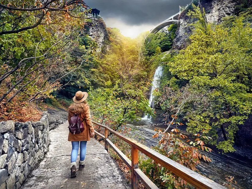 Şapkalı bir turist kadın, Gürcistan'ın Tiflis şehrinde, bulutlu bir havada, sonbahar ağaçlarının bulunduğu botanik bahçesindeki şelaleye doğru gidiyor.