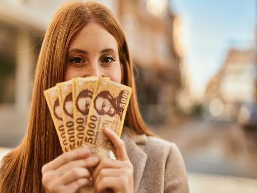 Genç kızıl saçlı kız, şehirde Macar forinti banknotlarıyla yüzünü kapatarak mutlu bir şekilde gülümsüyor.