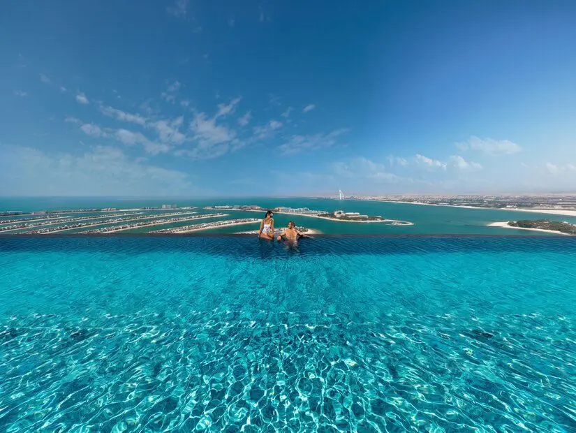 Dubai Palm Jumeirah manzaralı lüks sonsuzluk havuzunda (infinity pool) oturan ve manzaranın keyfini çıkaran mayolu bir çift, berrak turkuaz su ve geniş gökyüzü.
