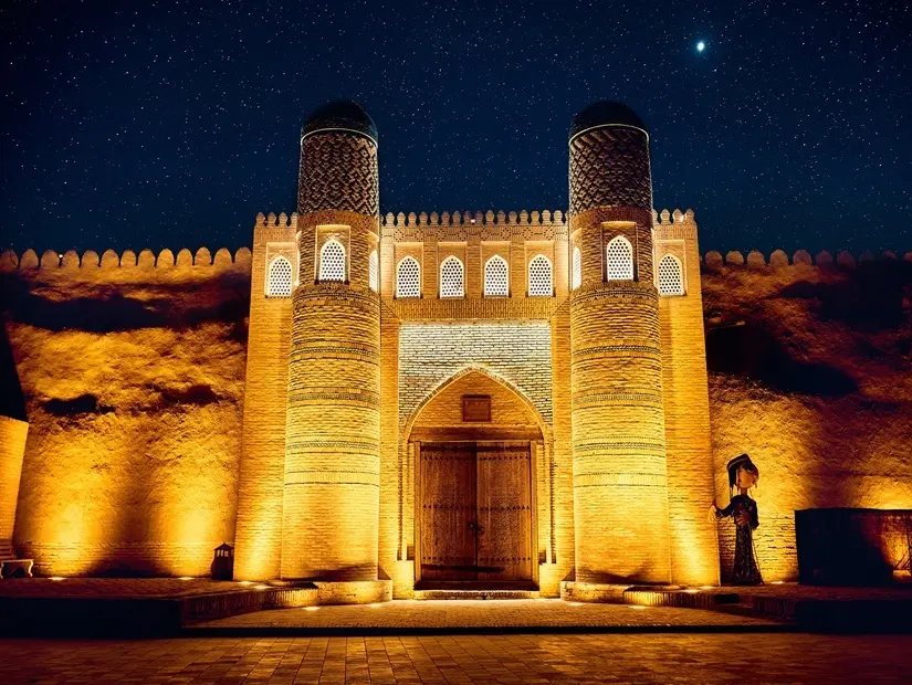 Özbekistan'ın Hiva kentindeki Itchan Kala şehrinde, gece gökyüzüne karşı yıldızlarla aydınlatılmış eski meydan ve kale kapısındaki Kalta Minor minaresinin tarihi mimarisi.