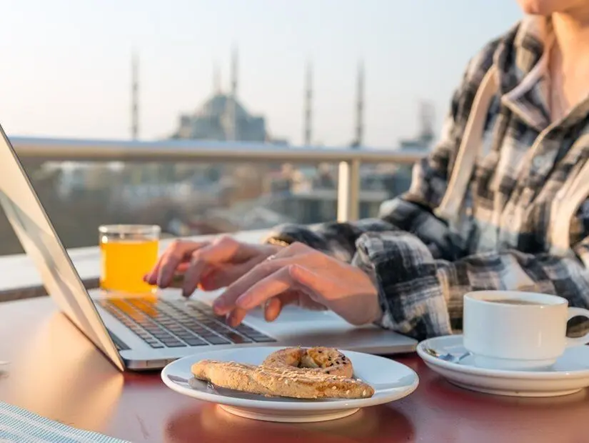 Kahvaltı yapan bir kişi, kafe terasında bir masada dizüstü bilgisayarında çalışırken, yanında kahve fincan görünümü